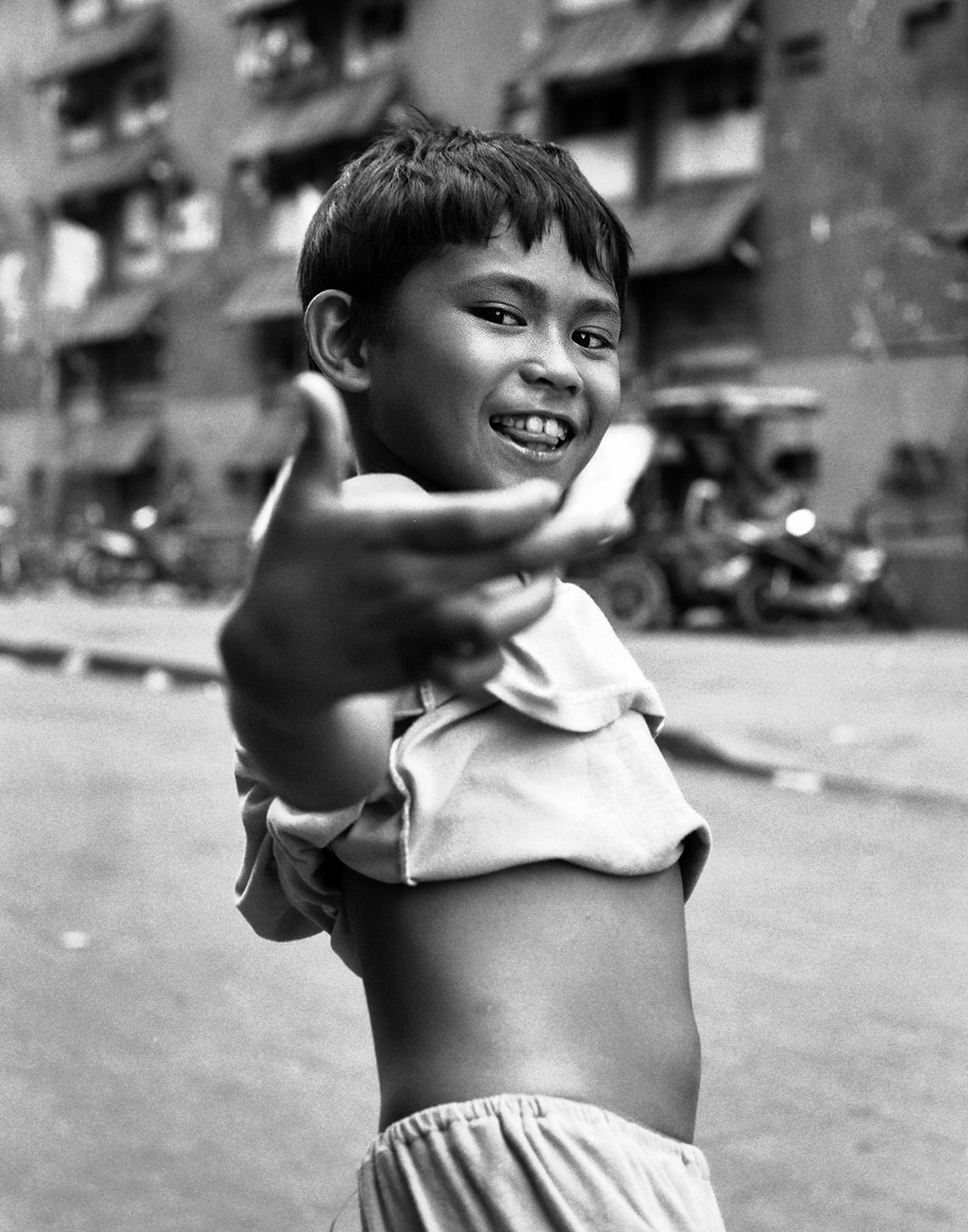 Young Filipino boy smiling and gesturing toward camera with outstretched hand wearing white shirt urban apartments in background