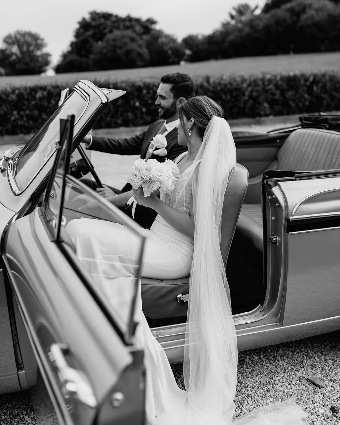 Black and white photo of a wedding couple sitting in a vintage convertible car, smiling at each other