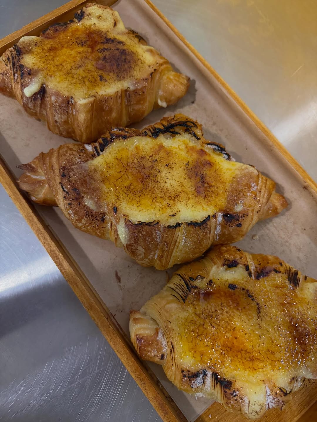Three crème brûlée croissants with caramelised golden tops on a wooden tray