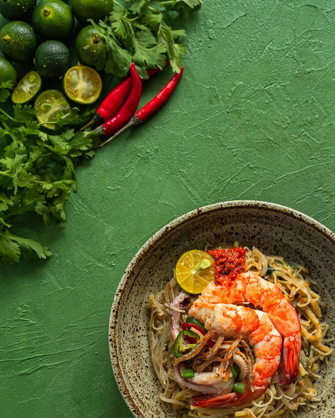 Hokkien Mee styled in a ceramic bowl on a green textured surface with fresh herbs and chilies