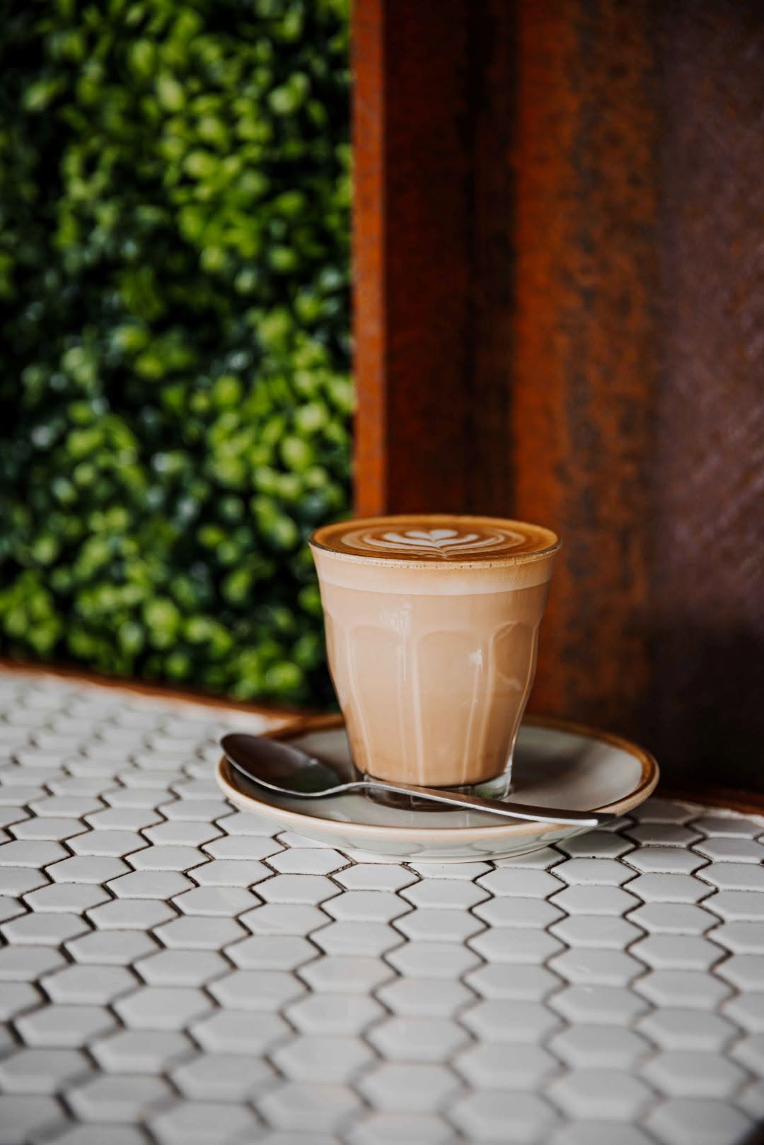 Specialty latte in glass cup on white hexagon tiles with green hedge backdrop
