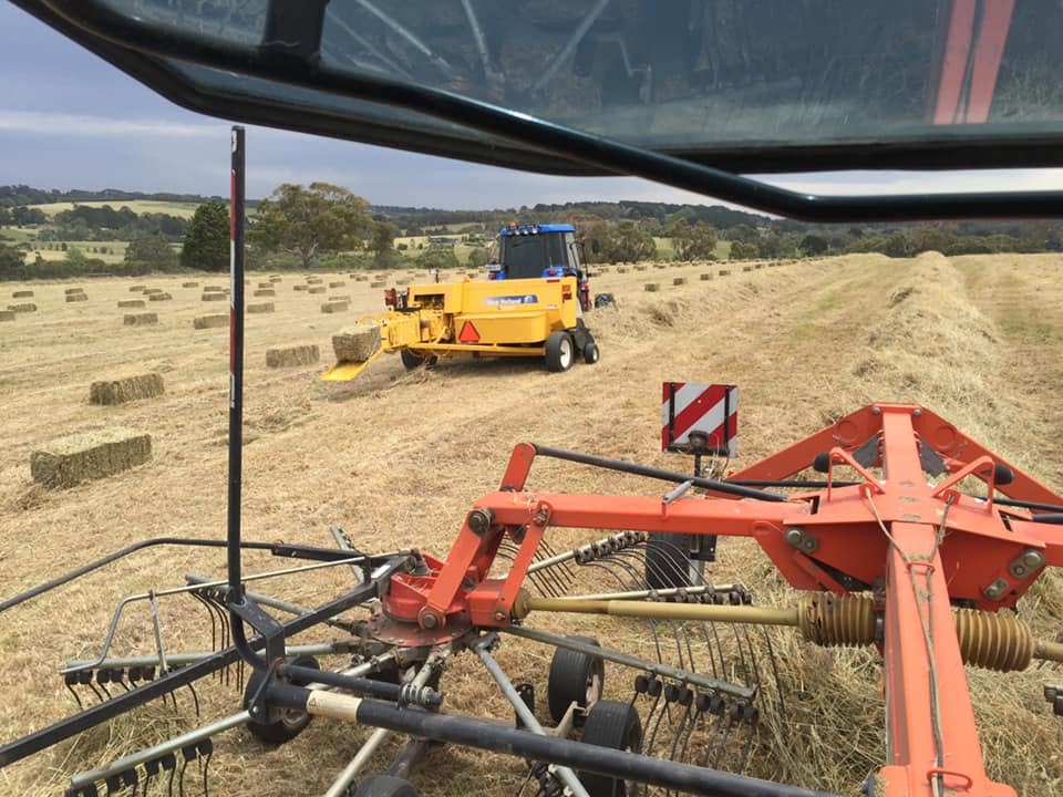 View from inside a tractor cab showing orange raking equipment in the foreground and a yellow baler working rows of cut hay across a paddock