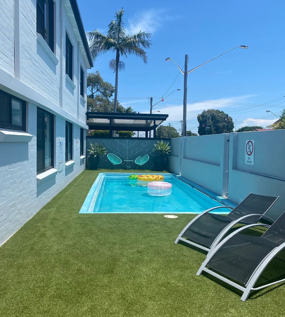 Homestead Motel exterior with outdoor pool, sun loungers, palm trees, and neatly manicured lawns under a blue sky