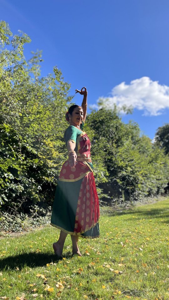 Dancer in green and red costume performing Bharatanatyam outdoors in garden with blue sky