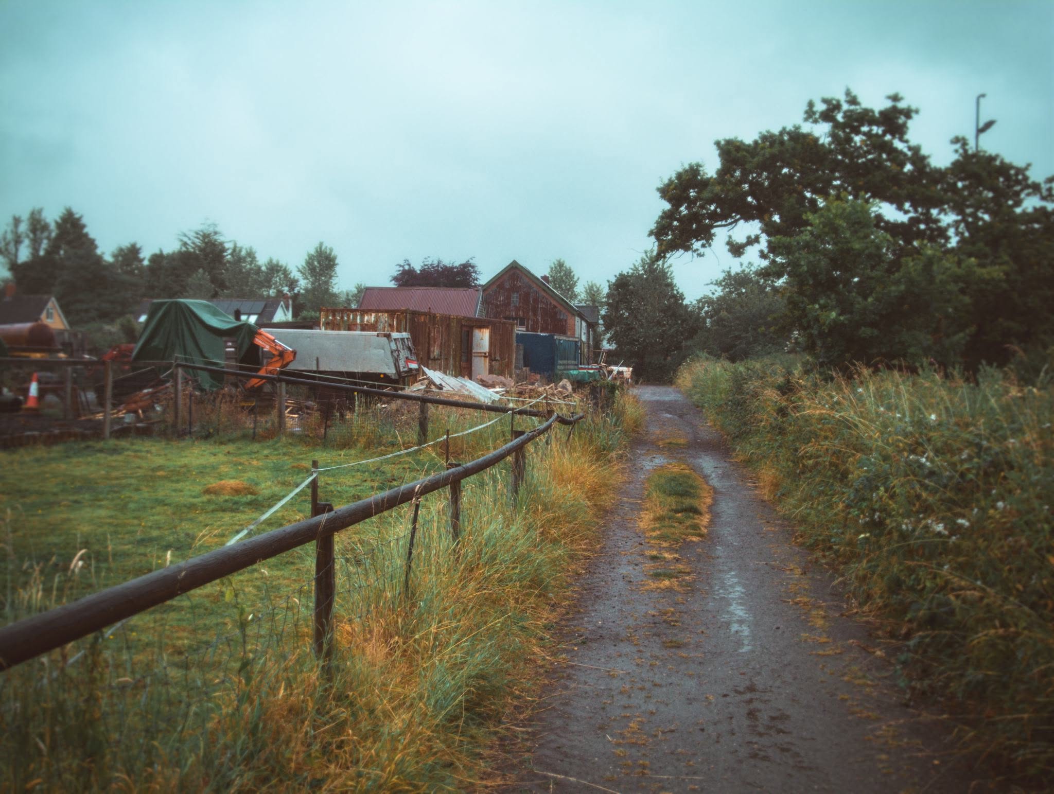 Colour photograph of a rural pathway with a wooden fence and farm buildings in the background