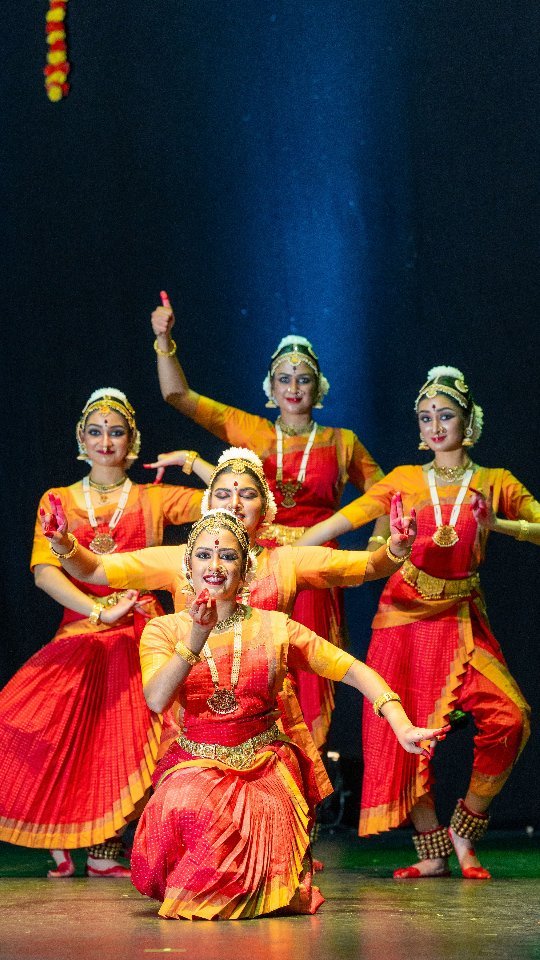 Group of five Bharatanatyam dancers in orange and red sarees performing synchronized movements on stage