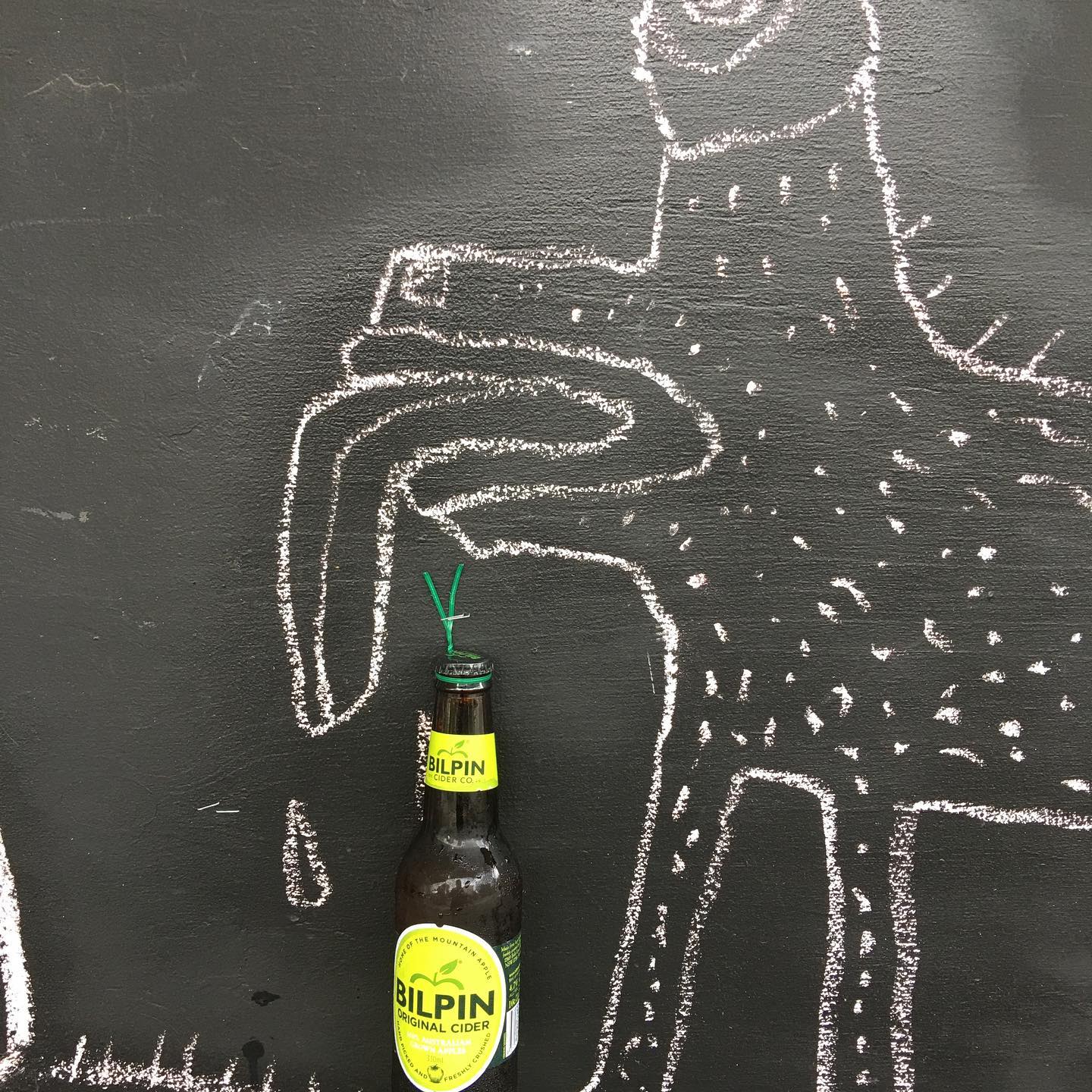 A single bottle of cider standing against a chalkboard wall with a chalk drawing