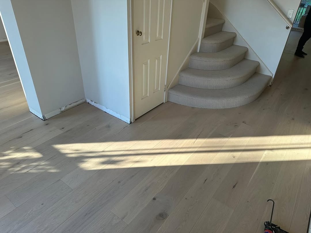 Wide-plank light oak flooring near a curved carpeted staircase base in a bright home