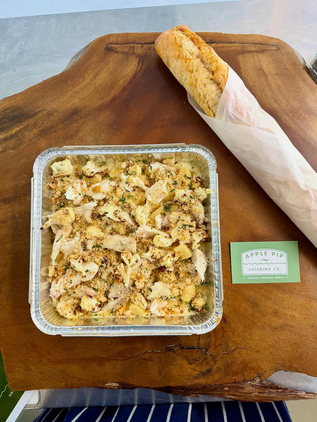 Fish pie in a foil tray with a crusty baguette on a timber board and Apple Pip Catering Co business card