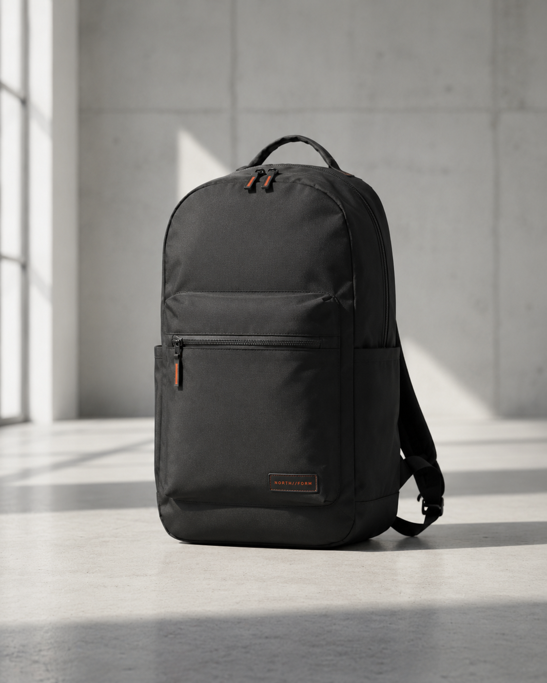Dark gray backpack with front zip pocket and dual orange zipper pulls in concrete studio
