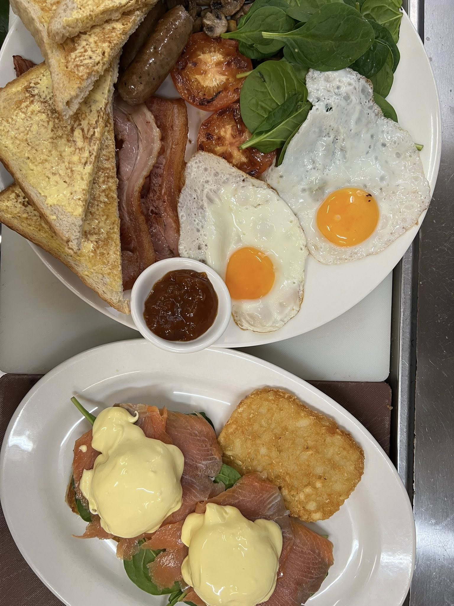 Big breakfast plate with two fried eggs, bacon, sausages, grilled tomatoes, spinach, mushrooms and toast alongside smoked salmon eggs benedict