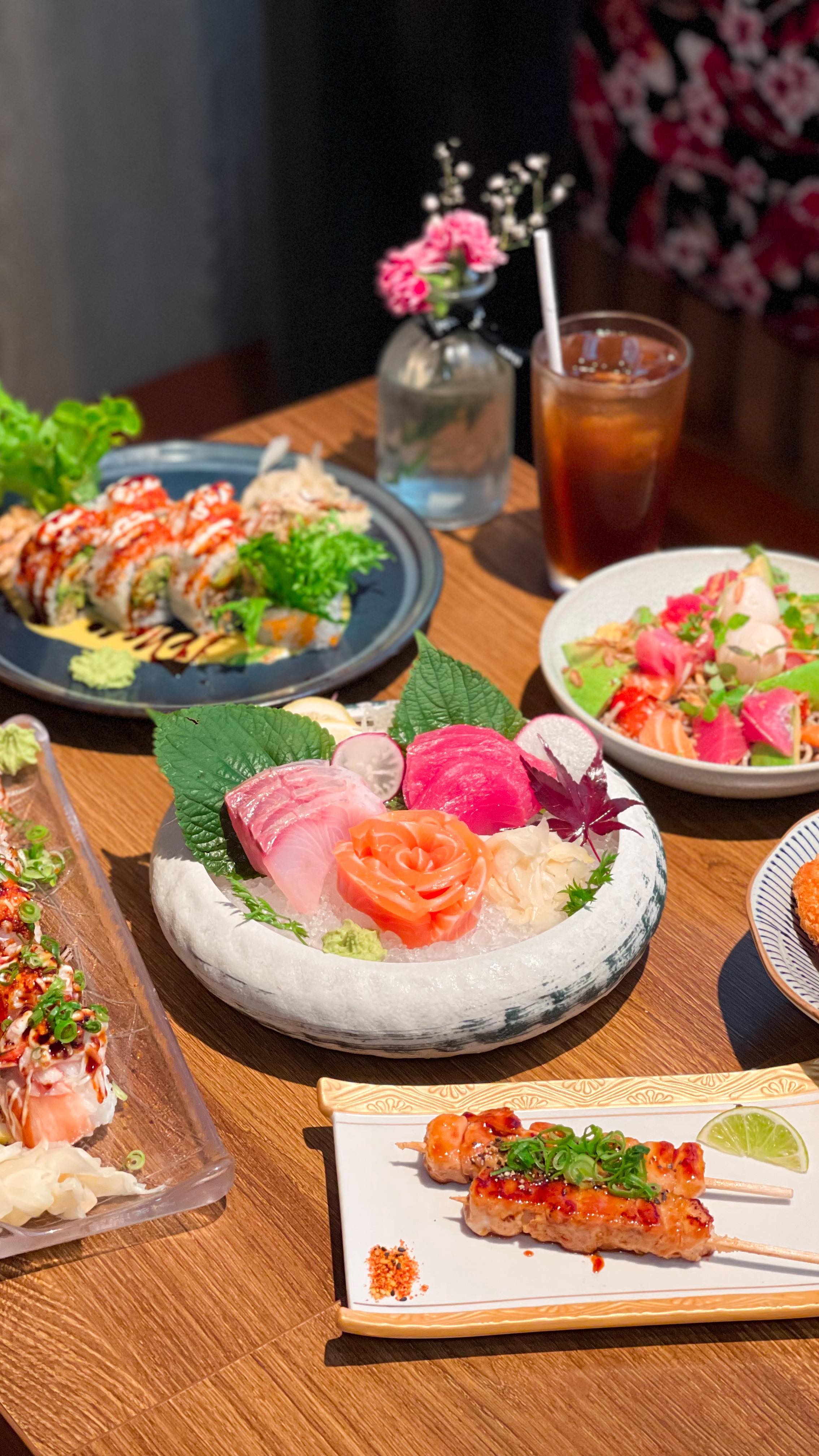 A spread of sushi rolls, sashimi, and grilled skewers on a wooden table at UMAYA World Square