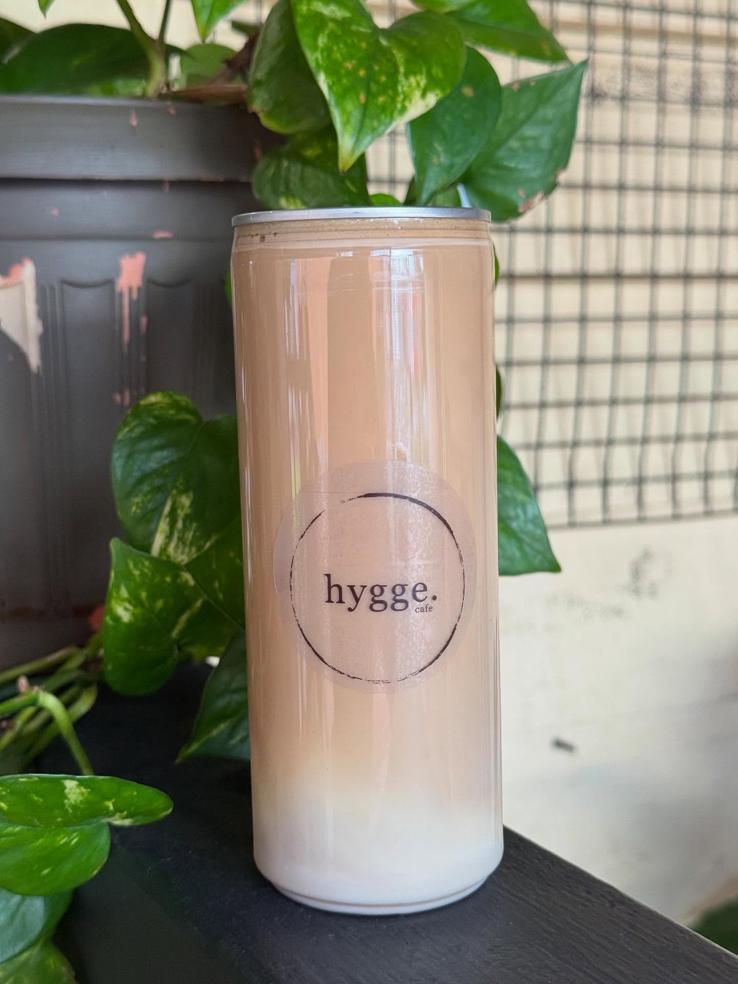 Hygge Cafe iced latte in a glass can with branded circle logo, surrounded by lush green plants