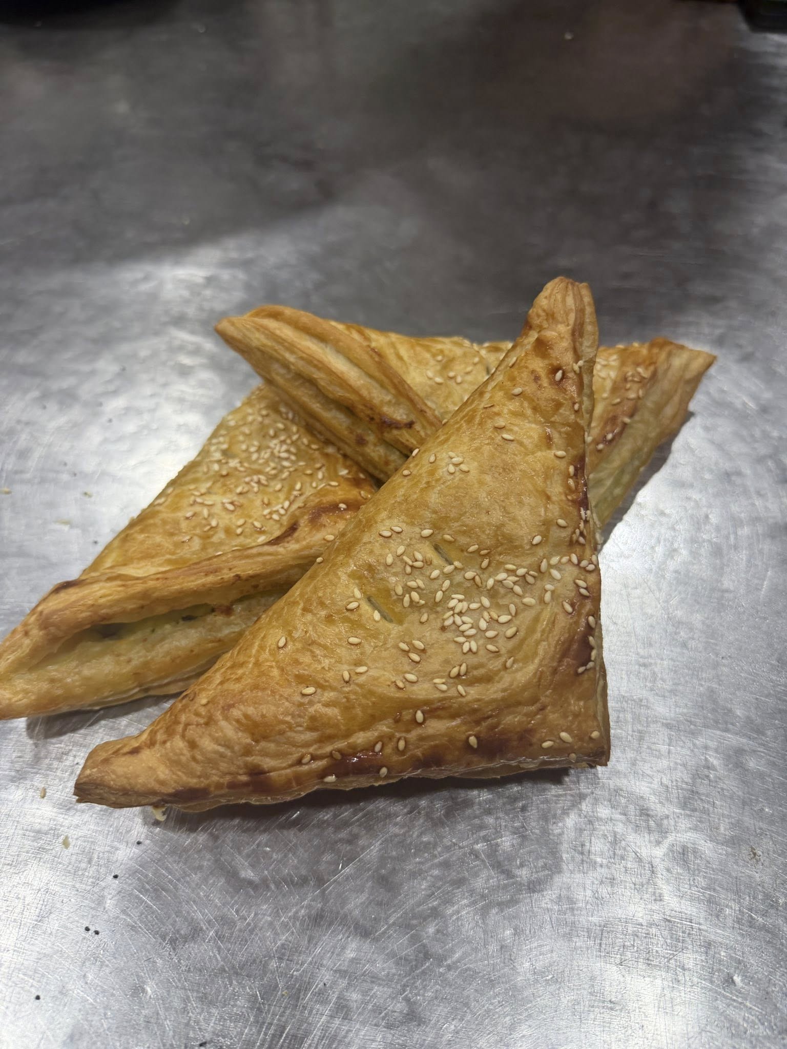Triangle-shaped puff pastry pockets with sesame seeds on a stainless steel bench
