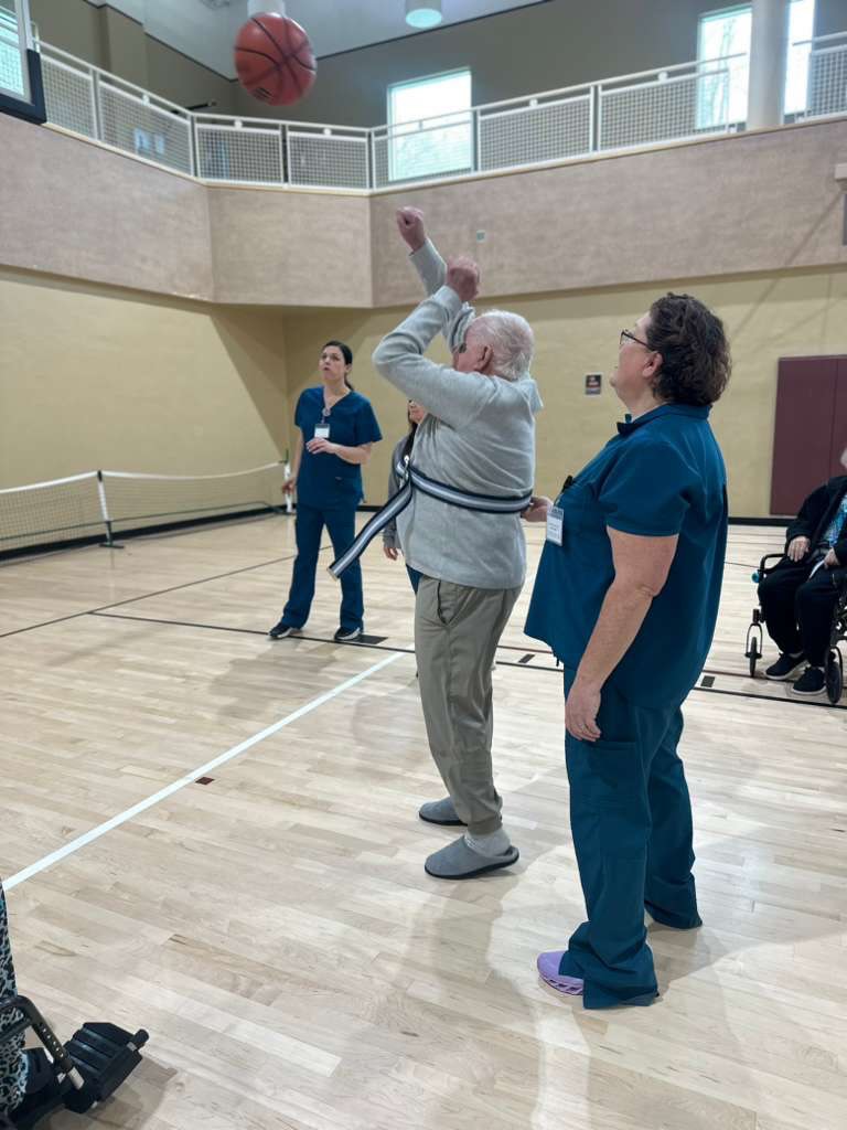 Therapy session in a gym with a resident shooting a basketball supported by a therapist