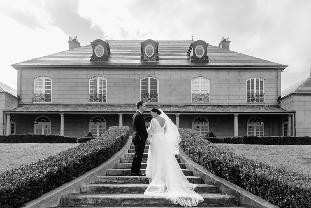 Black and white photograph of a bride and groom standing together on steps in front of a grand manor house