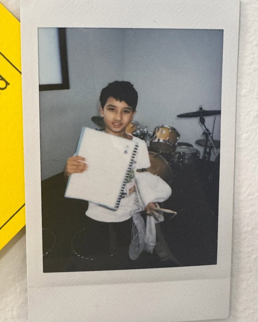 Child holding a music book in front of a drum kit