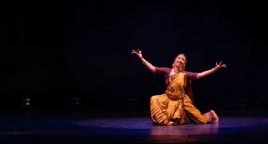 Solo Bharatanatyam dancer in golden-yellow costume performing on dark stage with arms extended