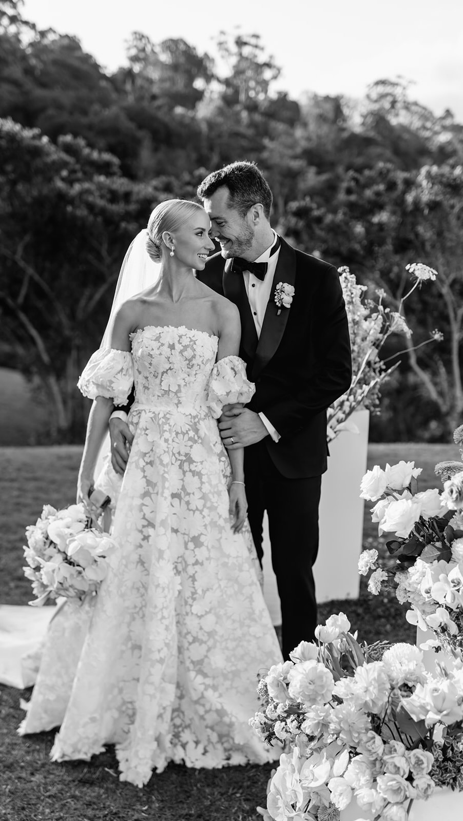 Black and white wedding ceremony photo — couple surrounded by lush white floral arrangements outdoors