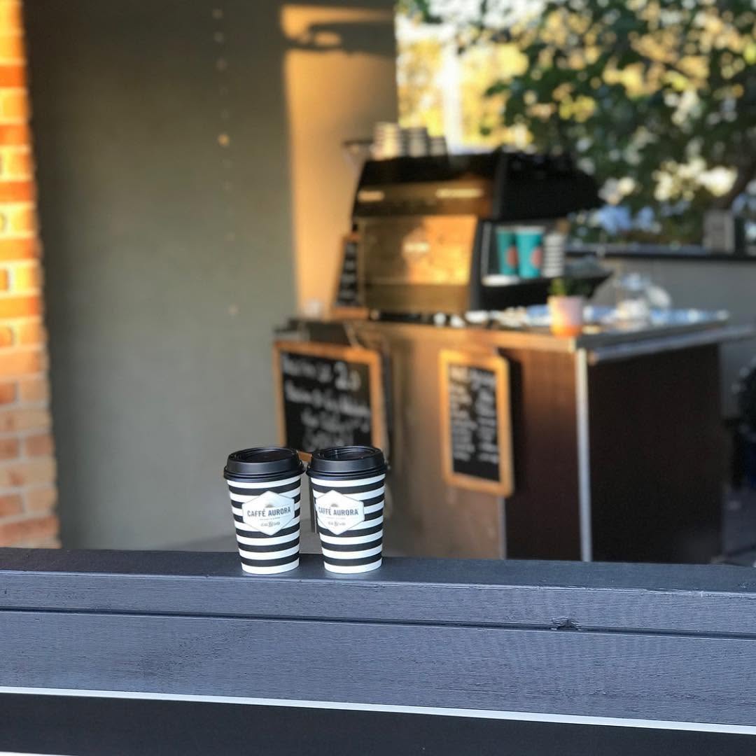 Two Caffè Aurora branded black-and-white striped takeaway coffee cups on a café counter ledge