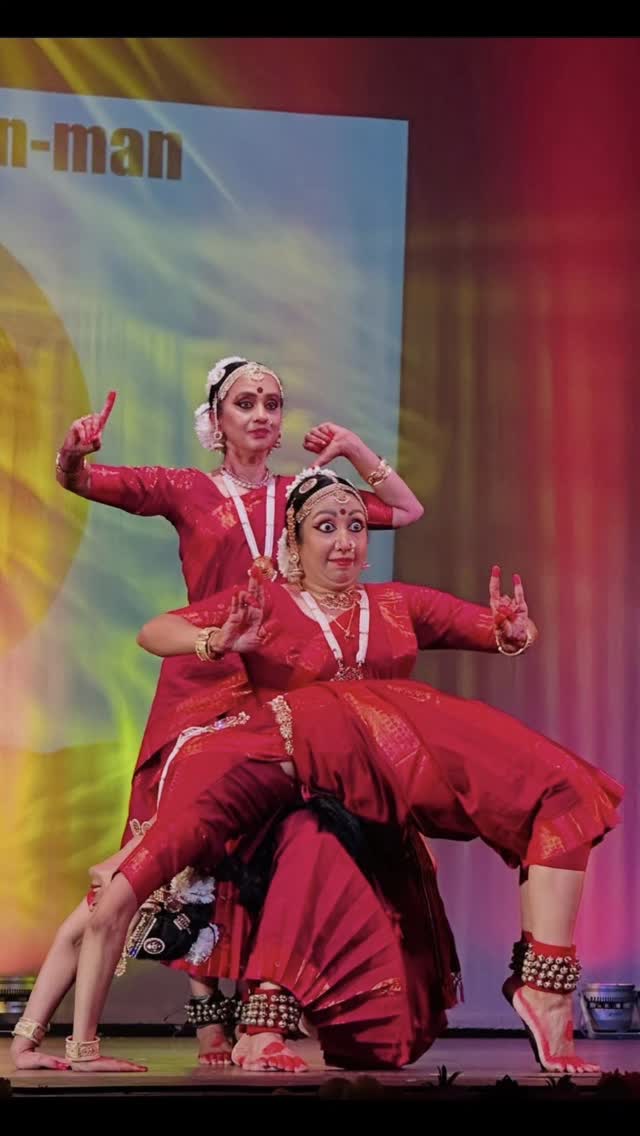 Two Bharatanatyam dancers in deep red sarees performing a dramatic pose on stage