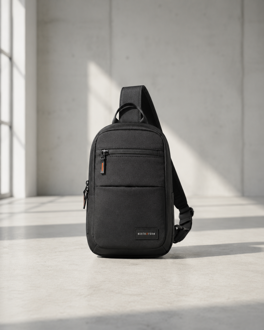 Black sling bag with single shoulder strap and orange zipper pull in concrete studio