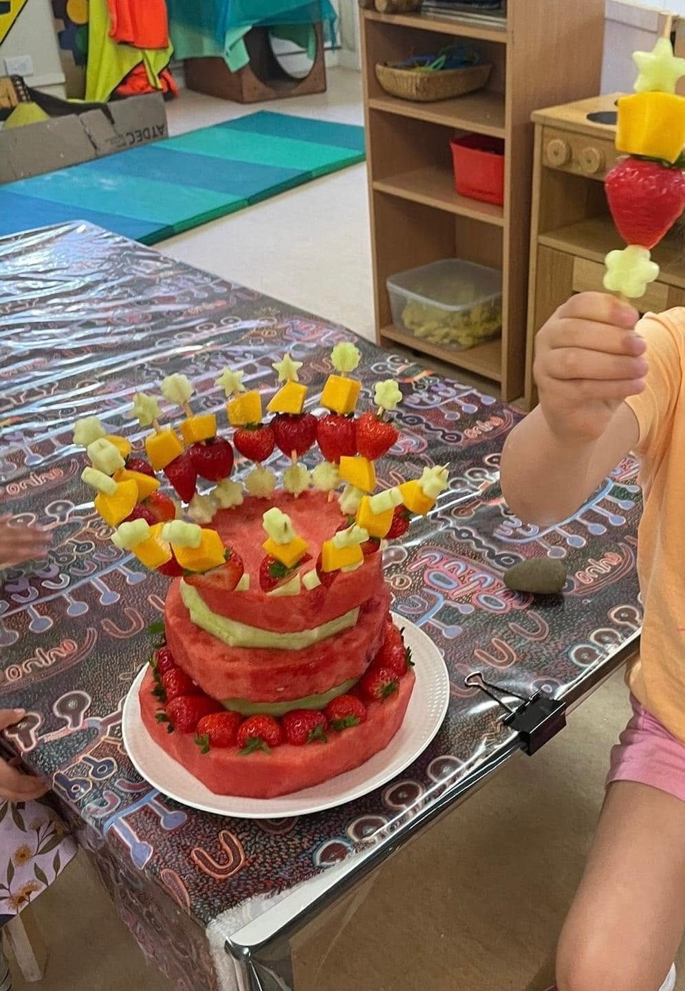 Colourful layered fruit cake decorated with strawberries, mango, and melon skewers — created during a healthy eating cooking activity at Dandenong North Preschool