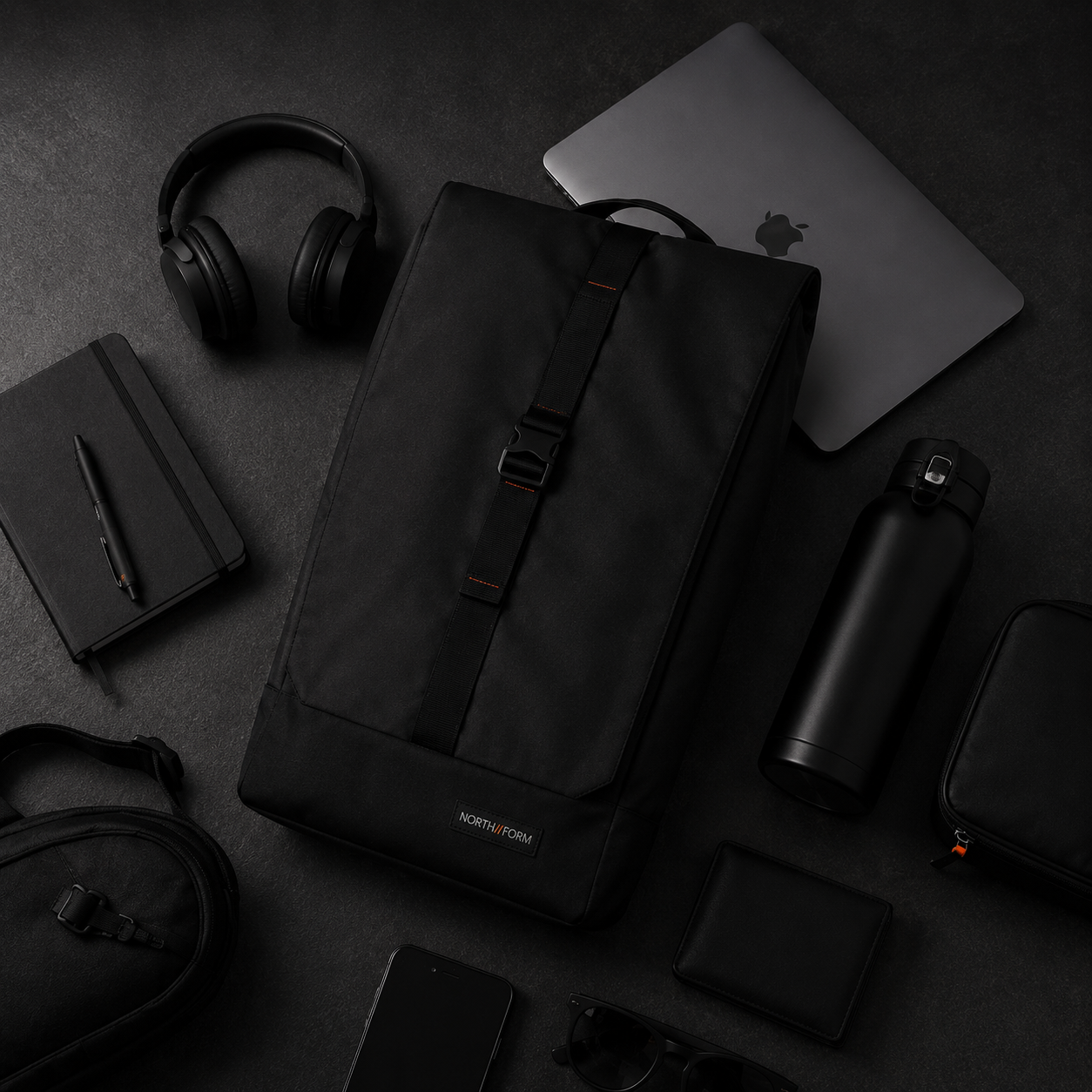 Flat lay of black NORTH//FORM backpack surrounded by everyday carry — headphones, MacBook, notebook, water bottle