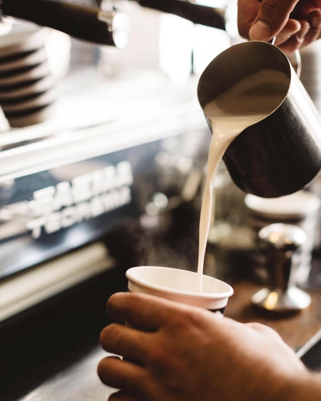 Close-up of steamed milk being poured from a silver jug into a takeaway cup at a Faema espresso machine