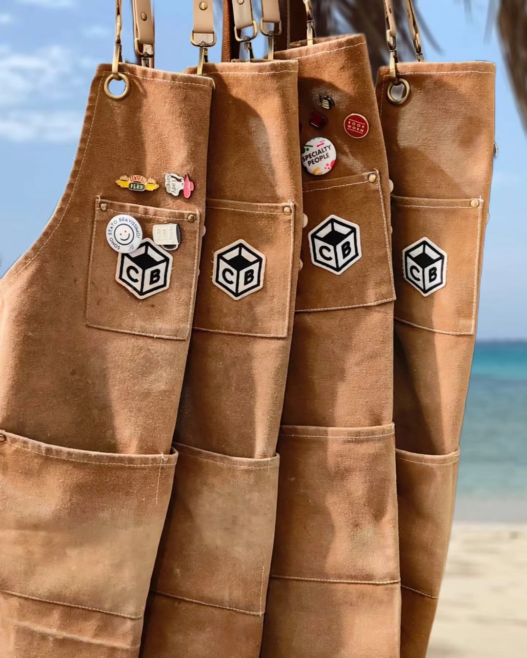 The Coffee Box branded barista aprons hanging at the beach — four canvas aprons with the CB logo patch and enamel pins