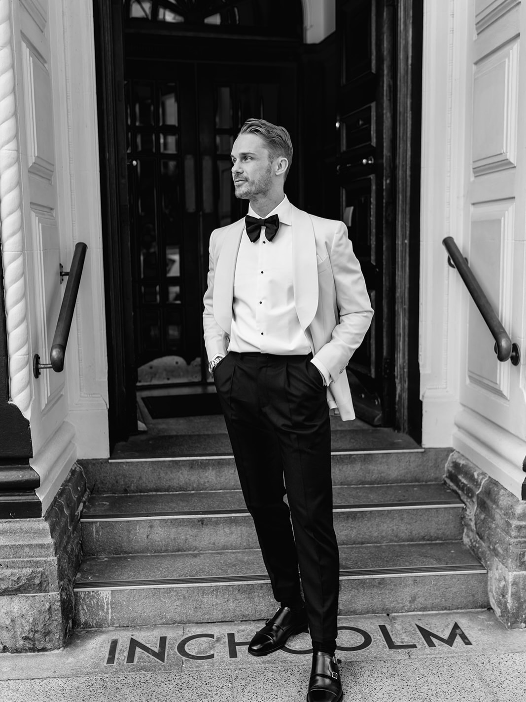 Black and white portrait of a groom standing on the steps of the Inchholm hotel in a white tuxedo