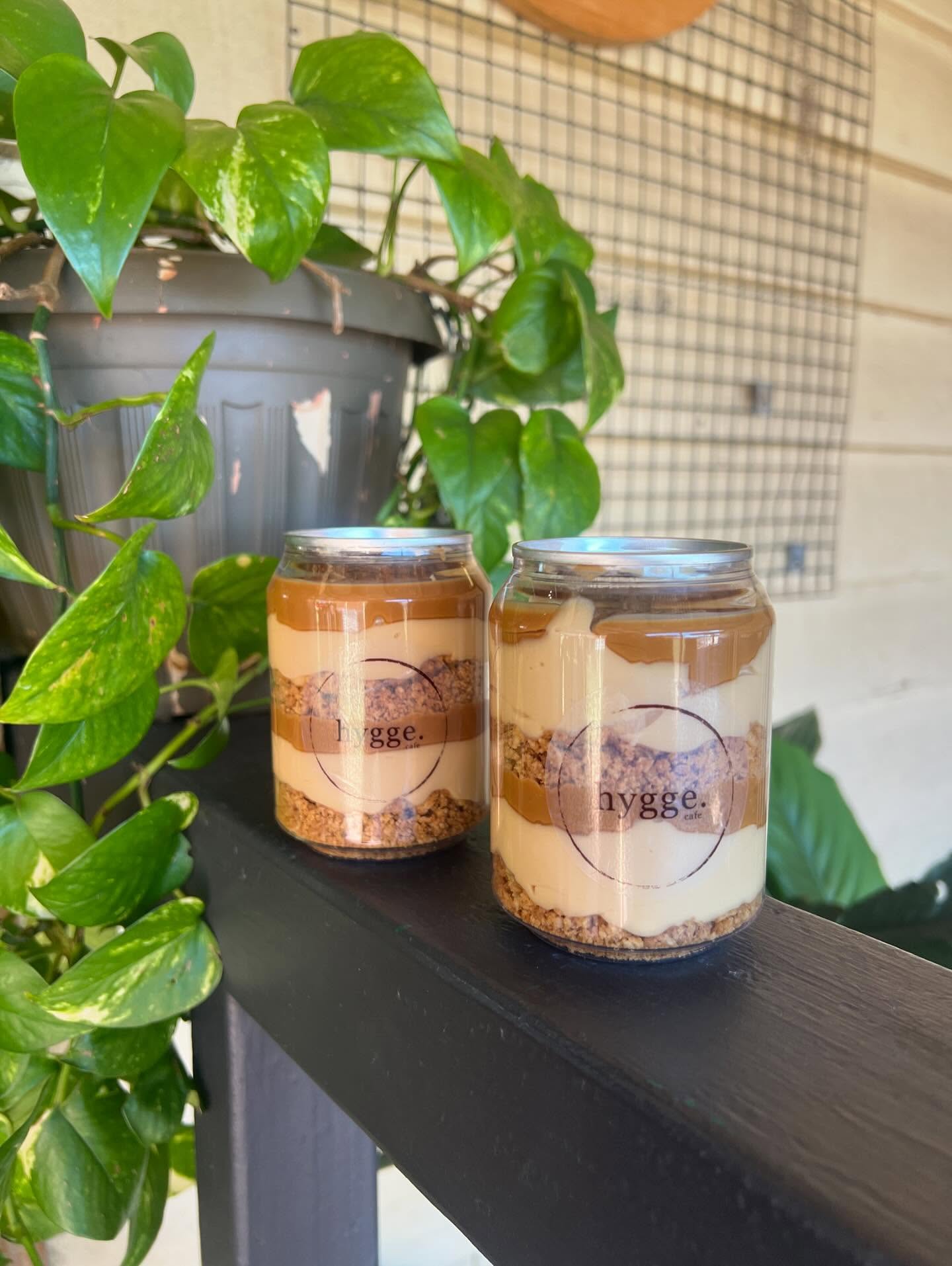 Two Hygge Cafe layered caramel dessert jars with branded circle logo, surrounded by green plants