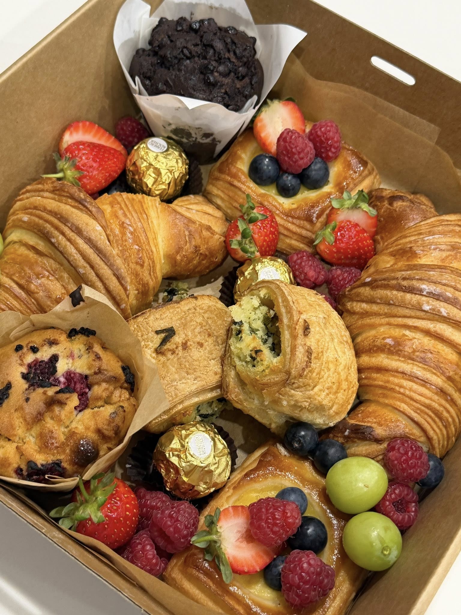 Open kraft box filled with croissants, a fruit Danish, blueberry muffin and fresh mixed berries