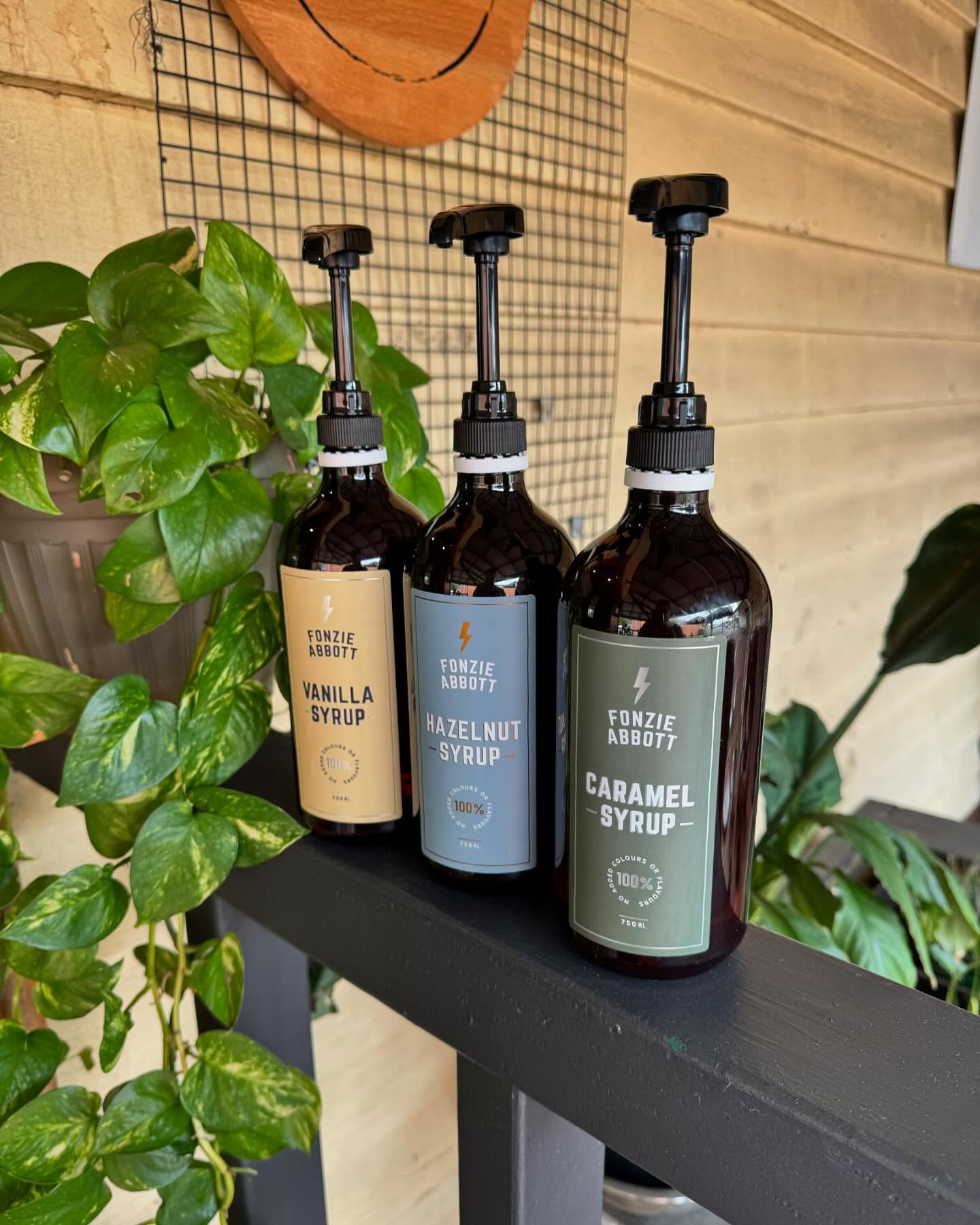 Three Fonzie Abbott flavour syrup bottles — vanilla, hazelnut and caramel — on a dark shelf with green foliage at Hygge Cafe