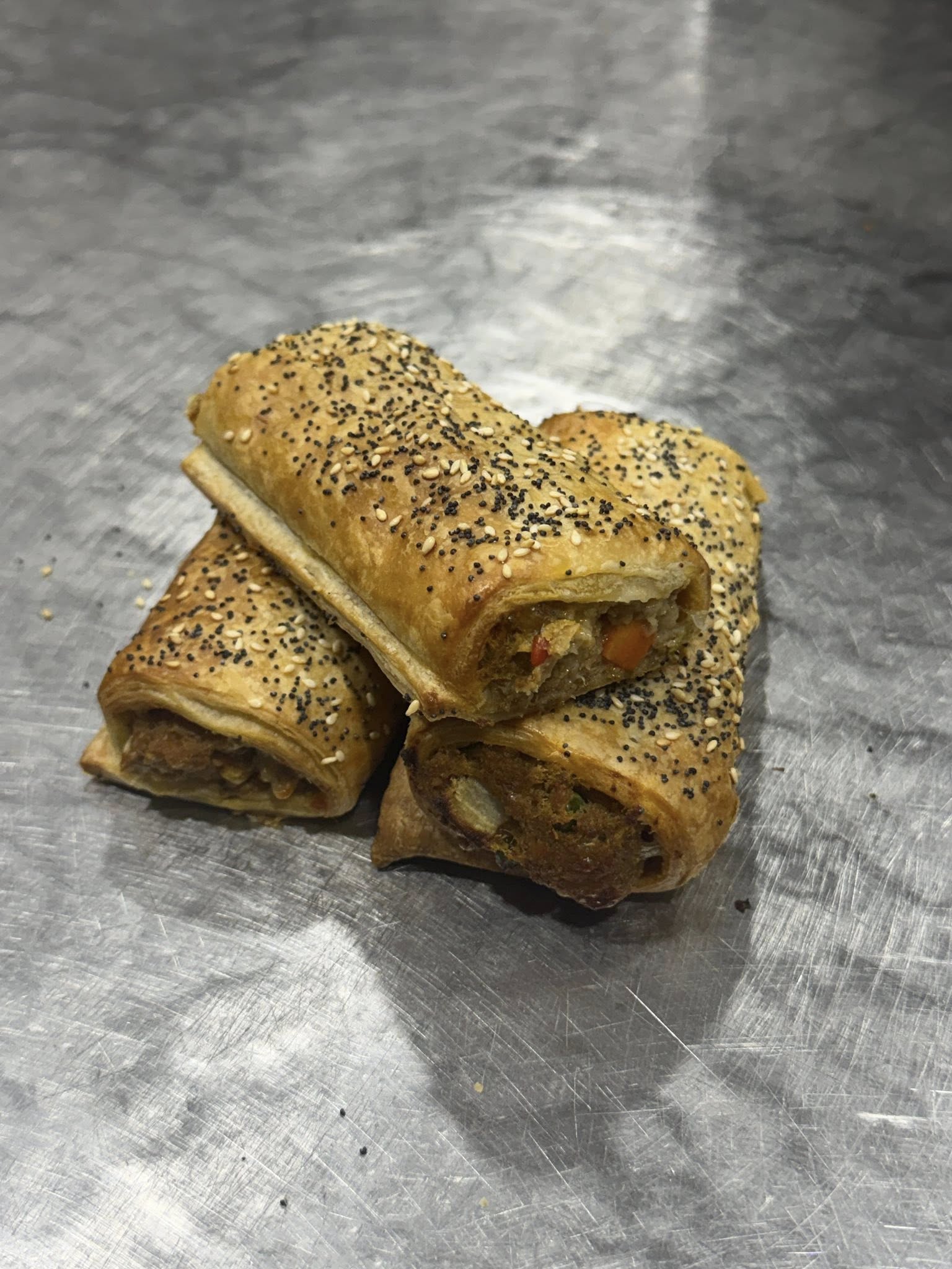 Two halves of a seeded sausage roll stacked on a stainless steel bench, showing the vegetable filling