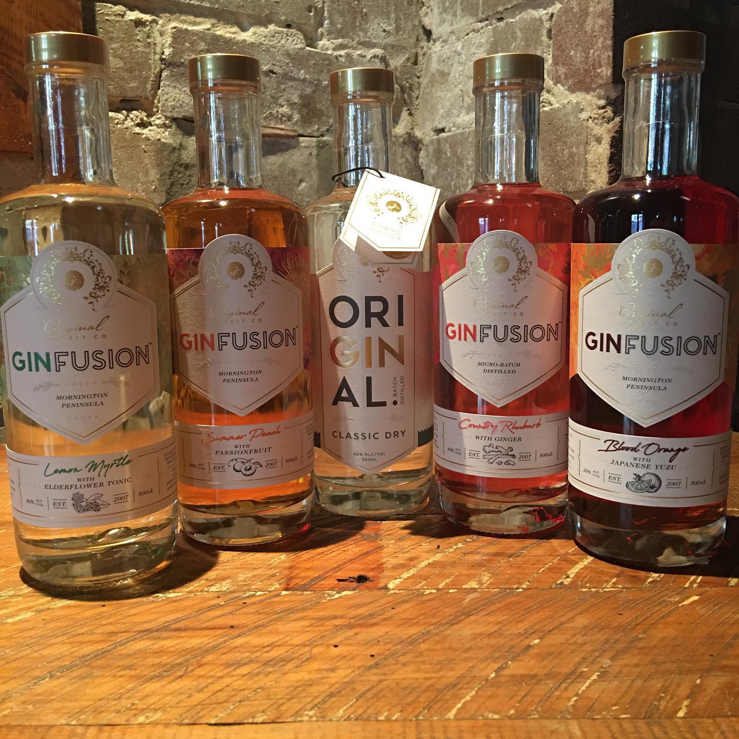 Five bottles of gin in varied colours lined up on a timber bench in front of a brick wall