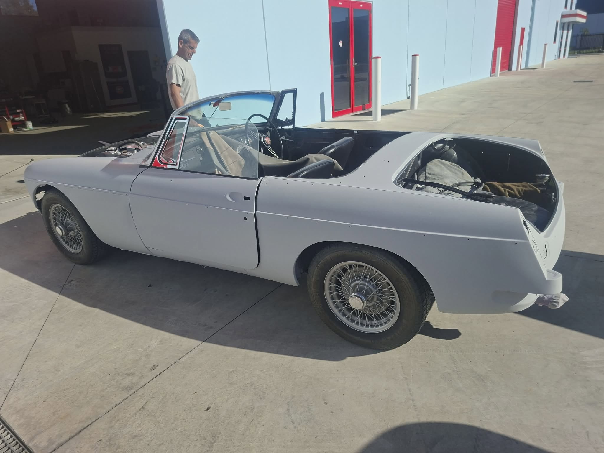 Classic MGB roadster in grey primer — rear quarter view outside the red-door workshop