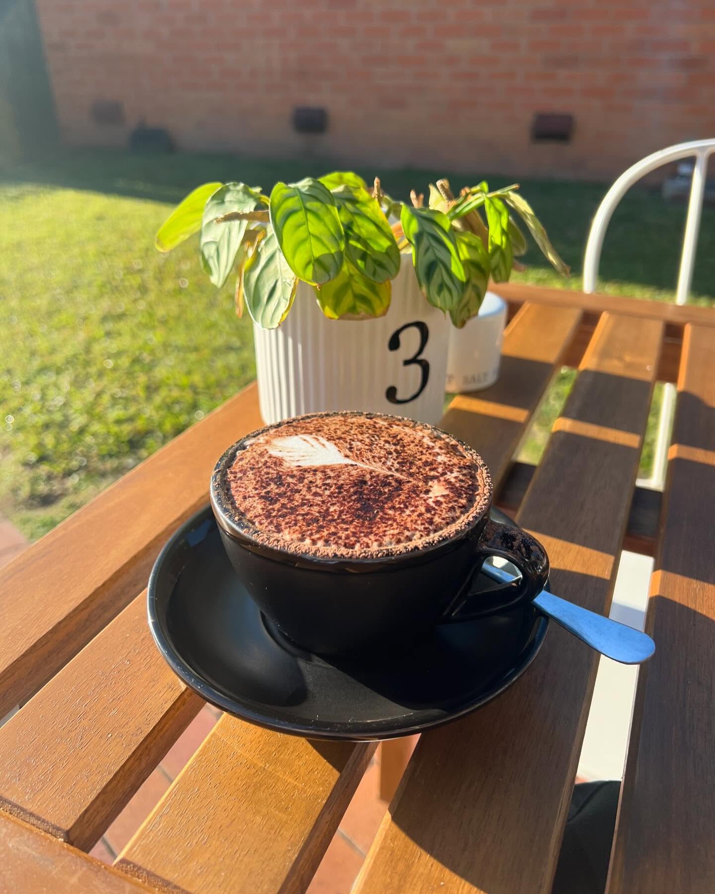 Hygge Cafe cappuccino with cocoa latte art in a black ceramic cup on a sunny timber table