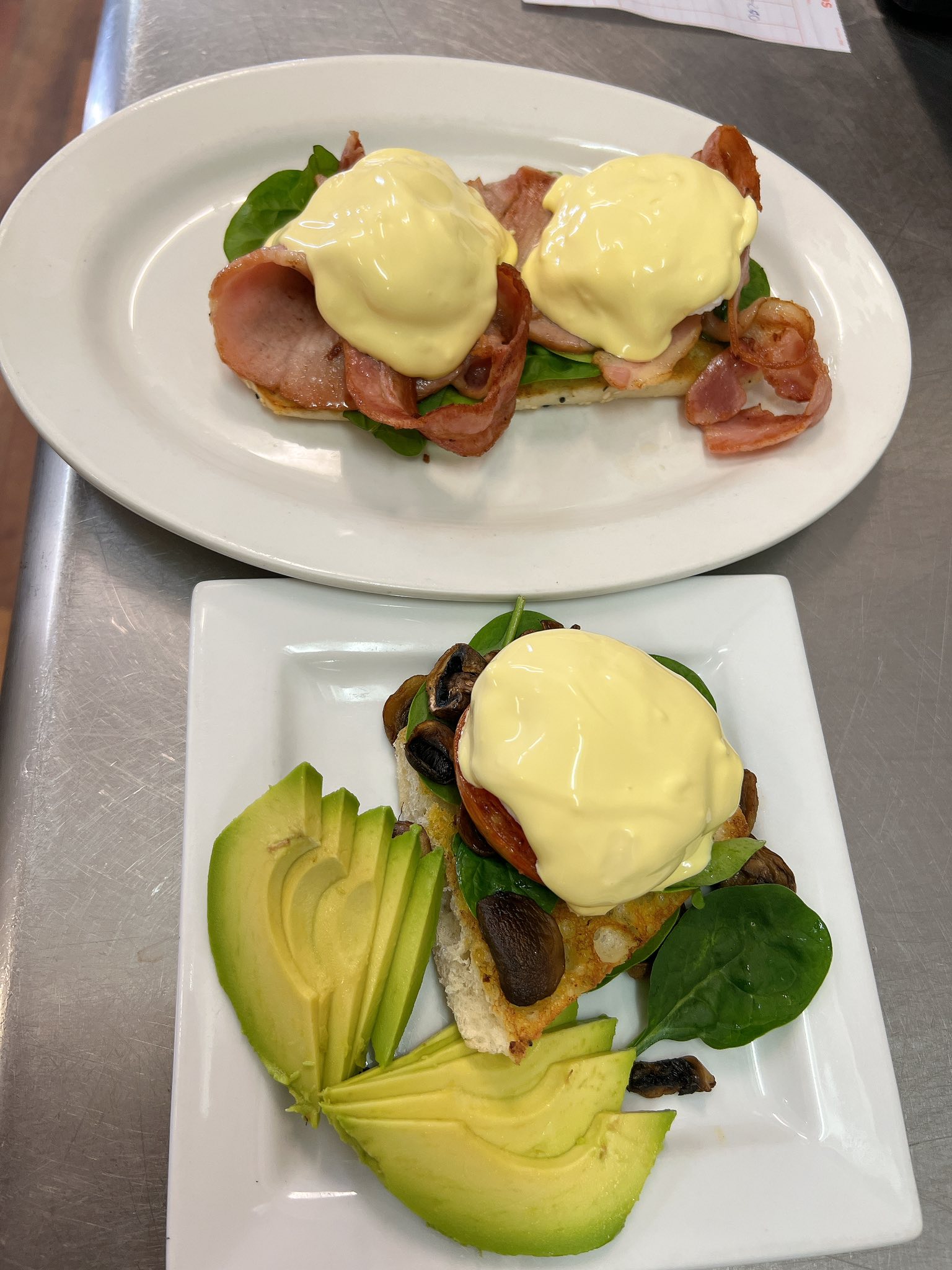 Two plates of eggs benedict — one with bacon hollandaise and one with mushrooms, avocado and hollandaise