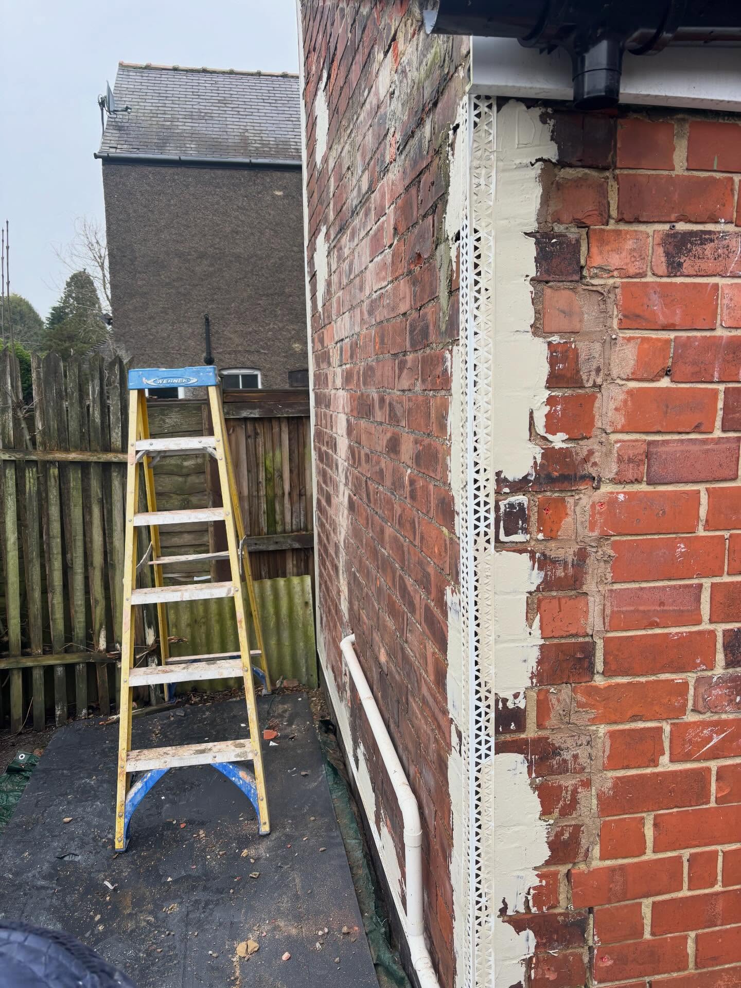Brick corner with metal corner bead and patches of cream plaster, with a ladder alongside
