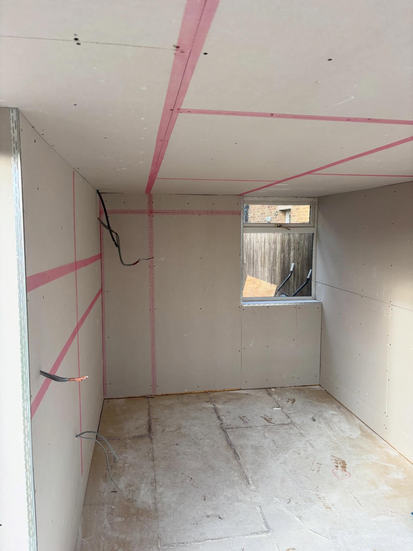 Room lined with plasterboard on walls and ceiling, joints taped in pink, with a small window