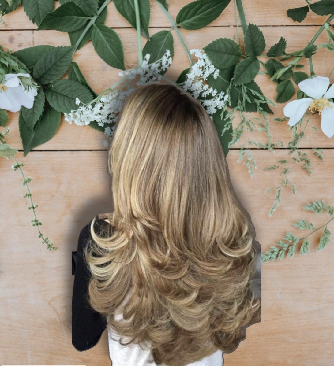 Long layered honey-blonde hair styled against a botanical backdrop of leaves and white flowers