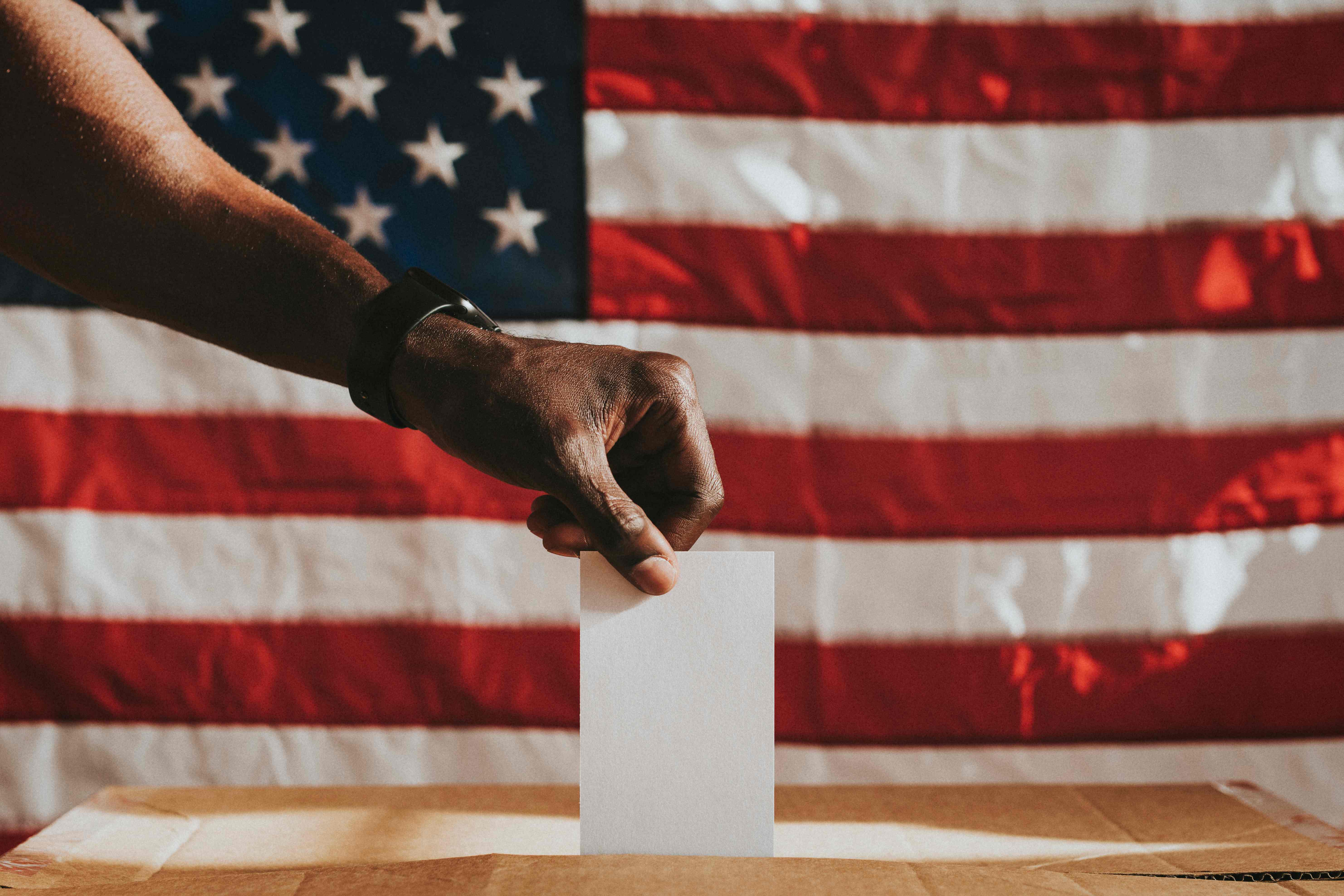 A hand placing a ballot into a box in front of an American flag