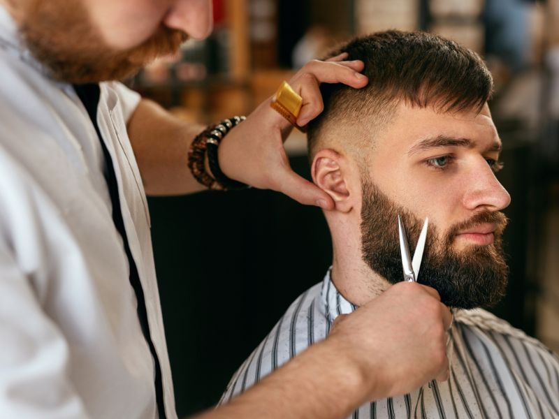 Barber trimming a bearded client’s hair and beard in a barbershop.
