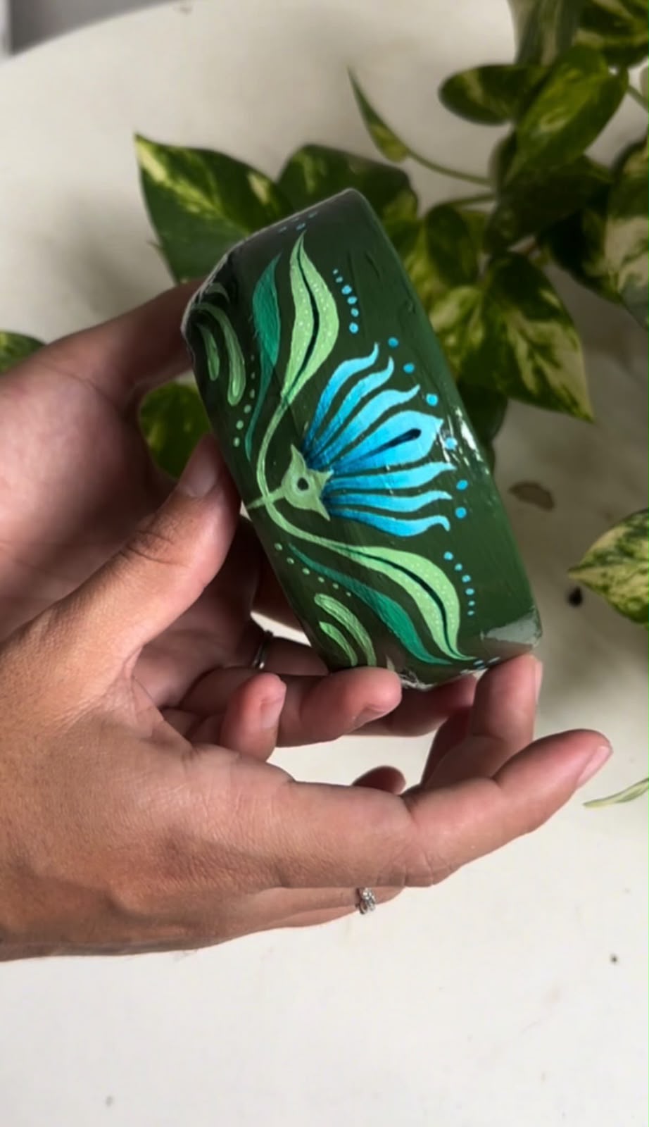 Potinho cerâmico verde escuro com borboleta e flor azul turquesa pintadas à mão, segurado na mão