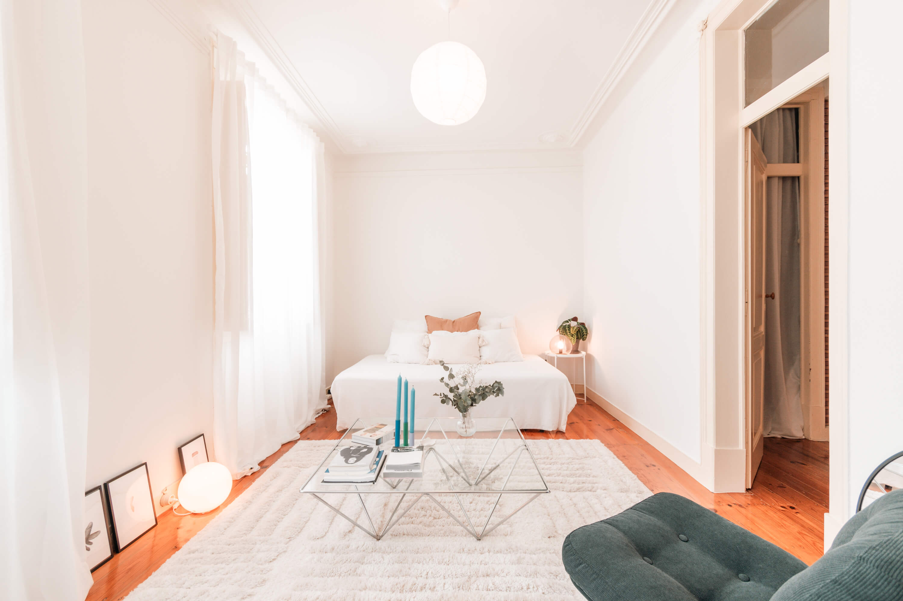 Minimalist living room with white walls, pine wood flooring, featuring a white bed, geometric glass coffee table, and teal accent chair