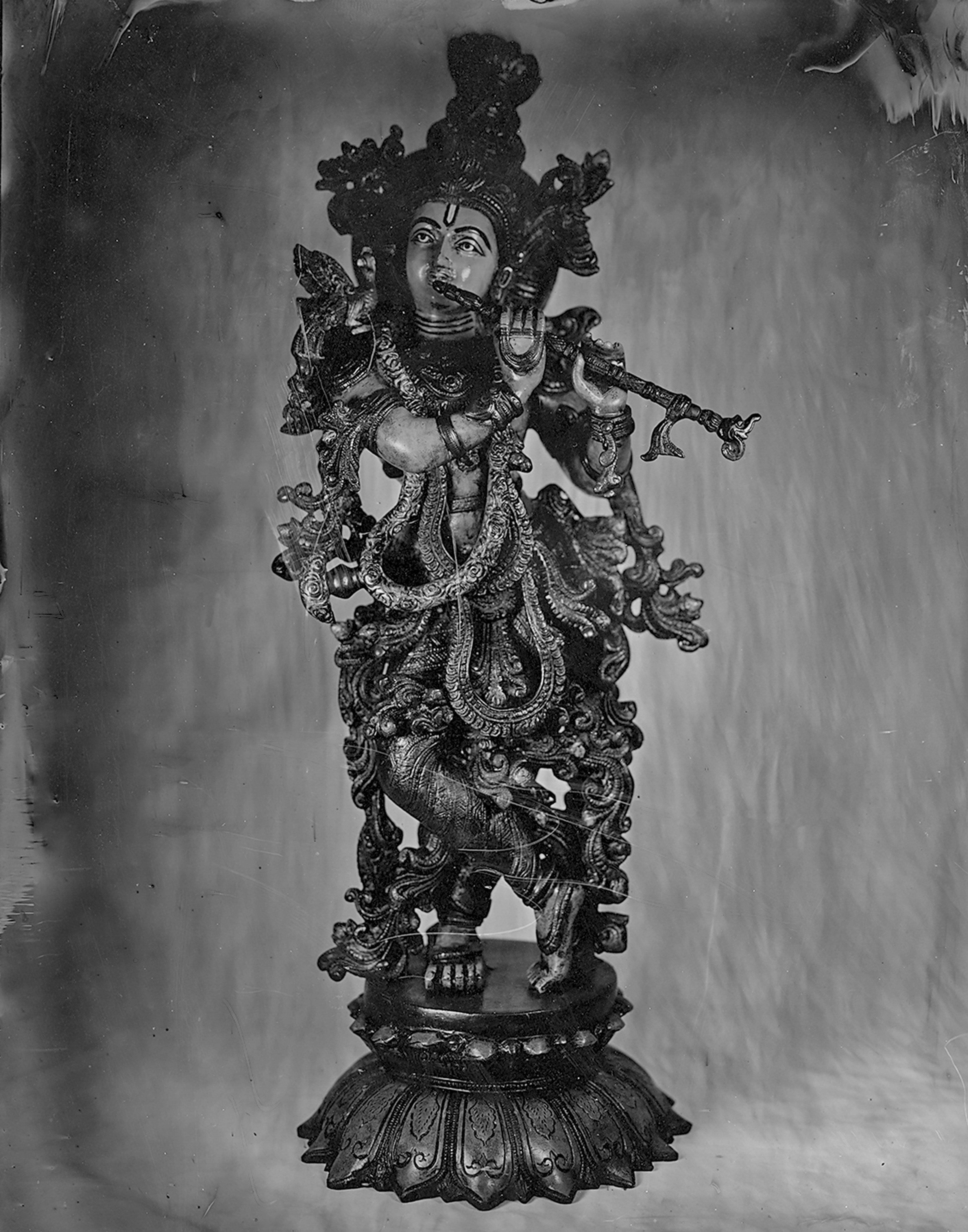 Ornate black and white photograph of elaborate Hindu deity statue with intricate sculptural details, multiple arms, decorative jewelry, and seated on ornamental lotus-base pedestal