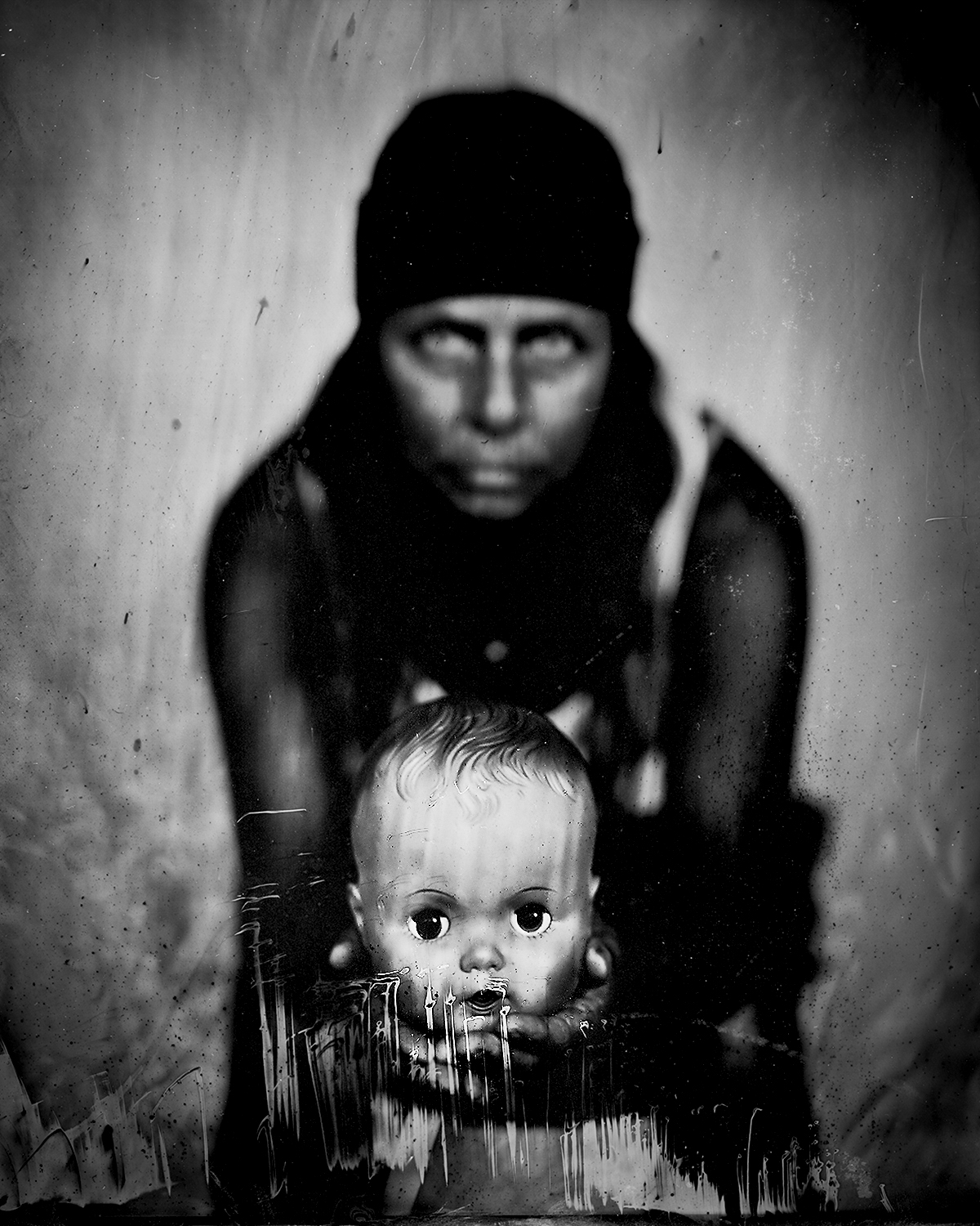Haunting collodion portrait of figure in dark beanie with obscured face looming behind vintage baby doll head with characteristic wet plate drips and chemical artifacts