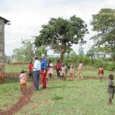 Besucher geht mit Dorfkindern auf einer grünen Wiese, Wassertank im Hintergrund