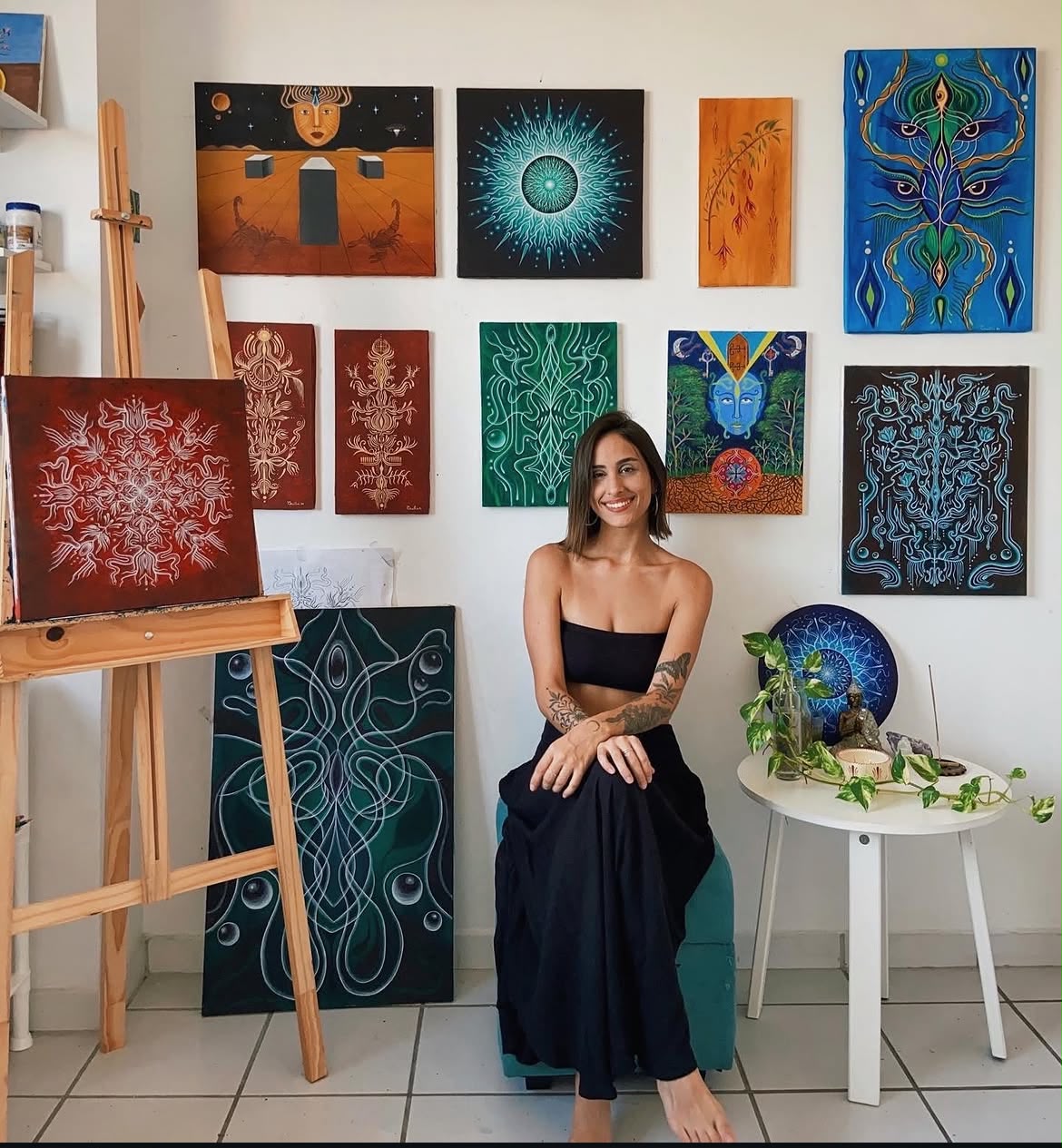 Cecília sentada no ateliê rodeada de pinturas originais nas paredes e cavaletes